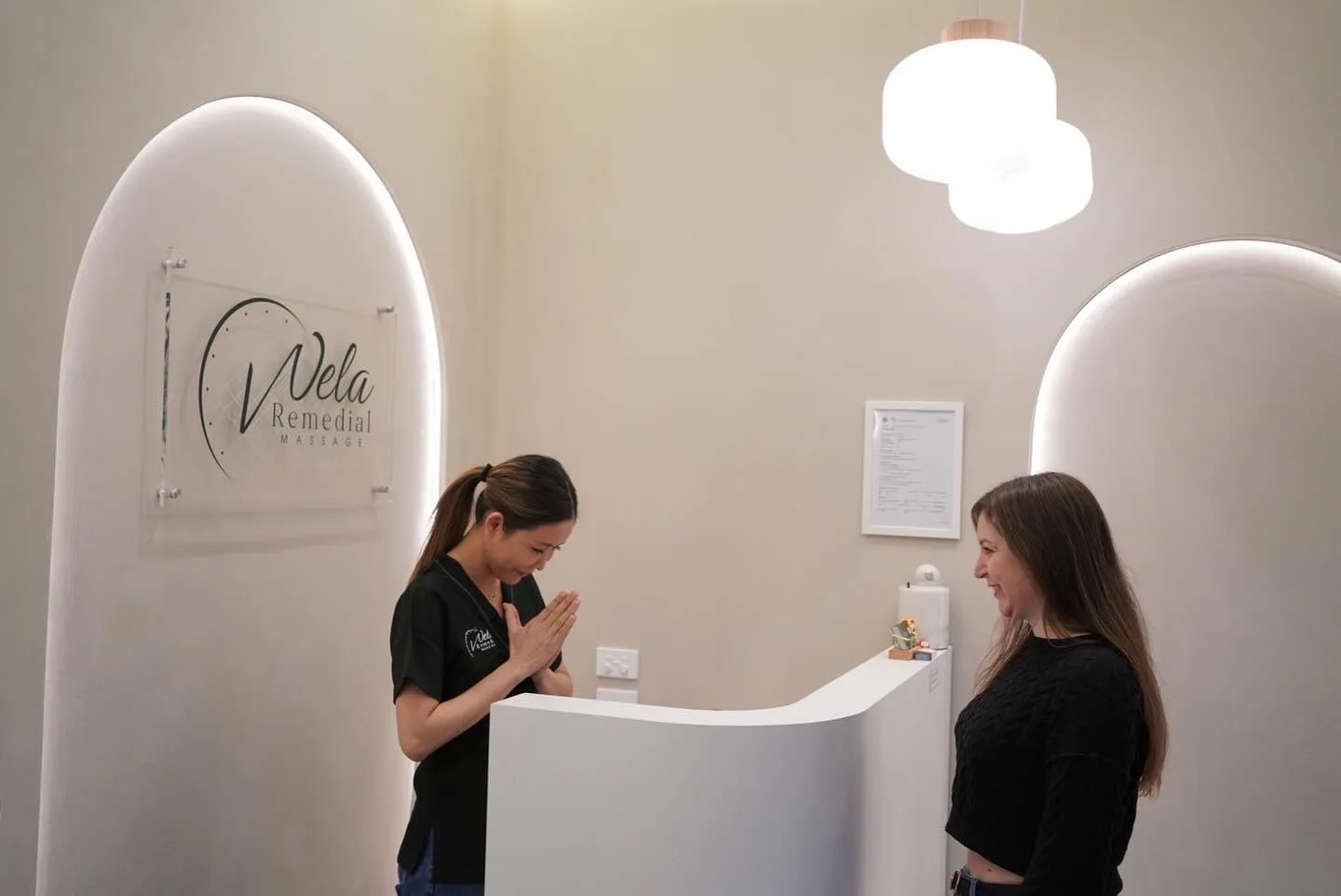 A Wela therapist greets a client with a warm wai bow across the curved reception desk, beneath the backlit gold Wela signage and twin pendant lamps